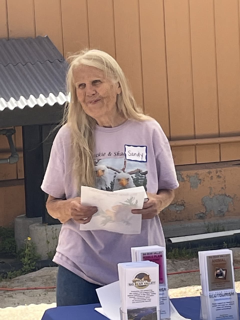 Sandy Steers behind an FOBBV outreach table, wearing a Jackie and Shadow tee with a handwritten name tag reading Sandy.